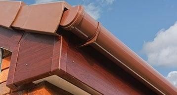 Close-up of a brown roof gutter system attached to the corner of a building, with a blue sky and white clouds in the background.