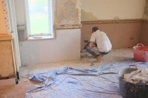 Person kneeling on the floor installing drywall in a partially renovated room, with tools and materials scattered around.