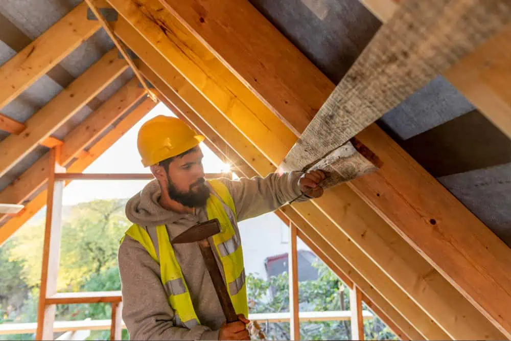 A construction worker wearing a hard hat and high-visibility vest uses a hammer and nails insulation to wooden beams inside an unfinished attic.